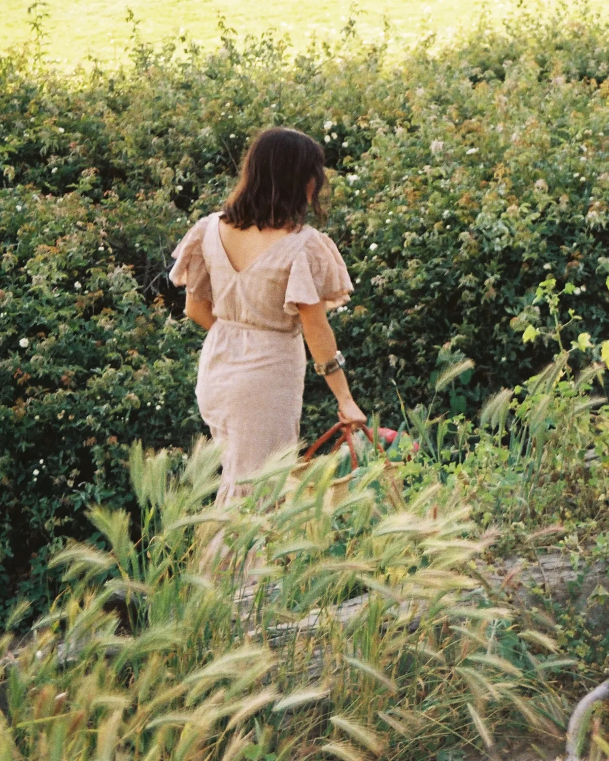 A woman in a soft blush dress walking through tall sunlit grass, carrying a basket